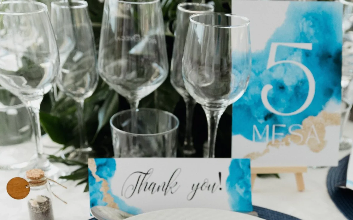 Detalle de la mesa: número de mesa 5 en acuarela azul y oro, tarjeta Thank you y botecito de cristal con arena y conchas