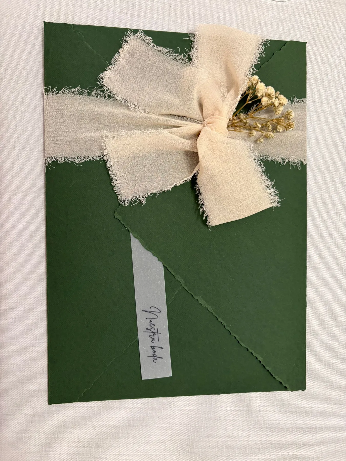 Sobre verde atado con cinta de gasa color crema, flores secas y una etiqueta de papel vegetal que dice Nuestra boda