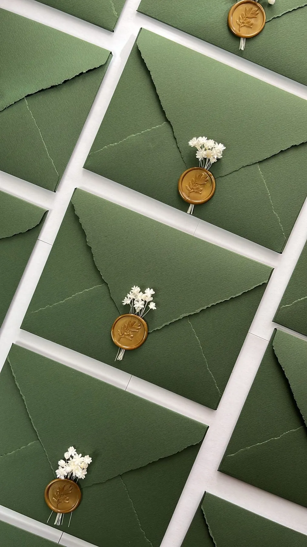 Sobres de papel verde con borde rasgado, cerrados con sello de lacre dorado y un ramillete de flores secas blancas
